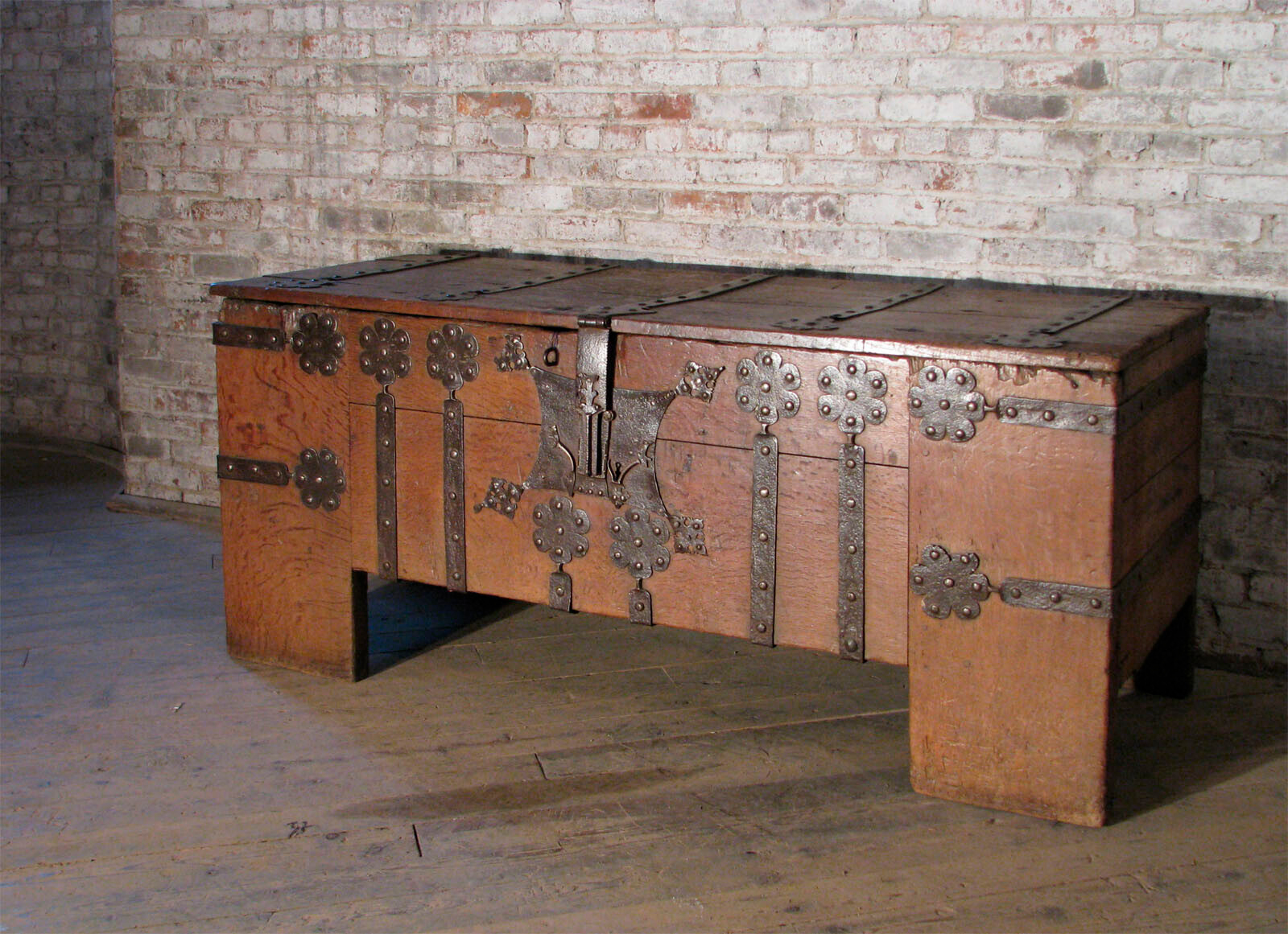 A large antique wooden chest with ornate metal hinges and decorations sits against a brick wall.