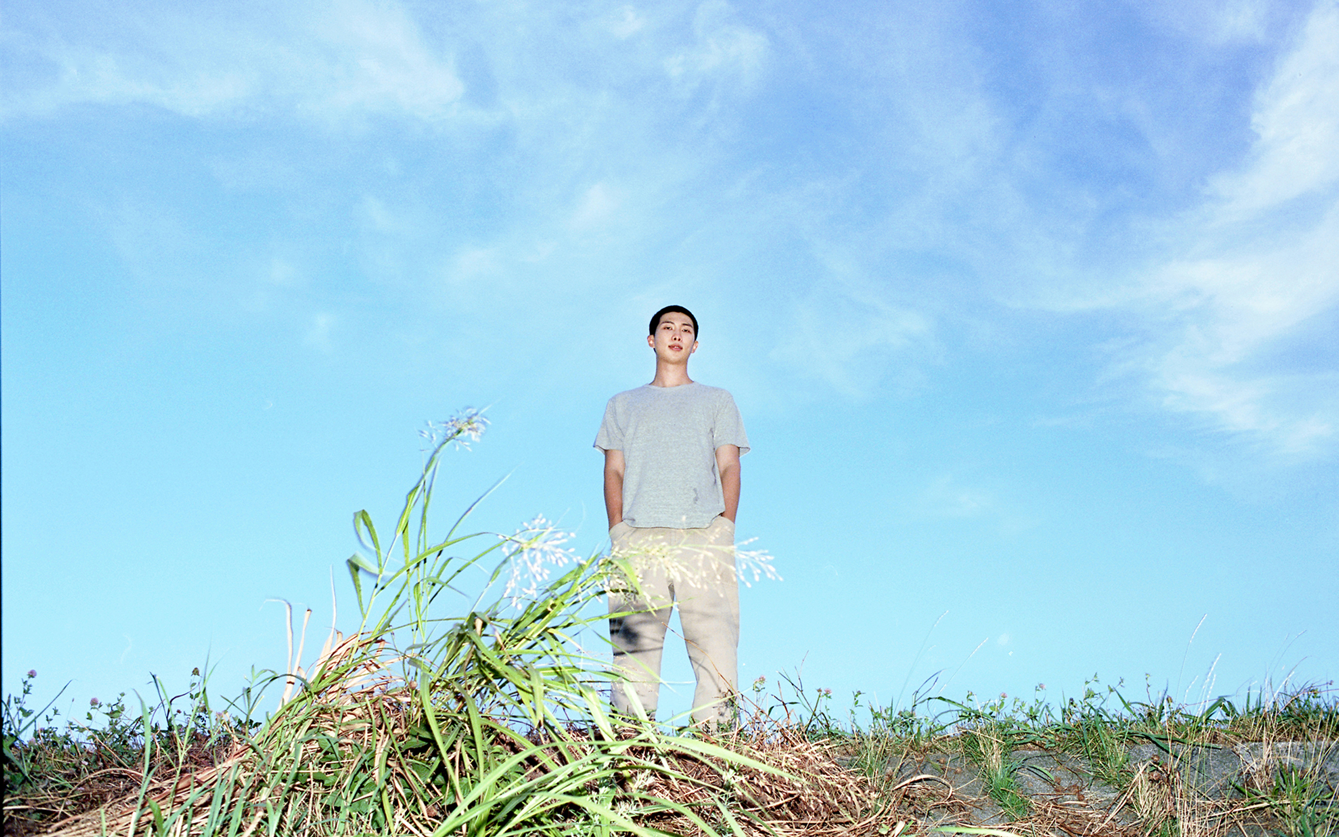 A man in a gray shirt stands on grass under a bright blue sky with light clouds.
