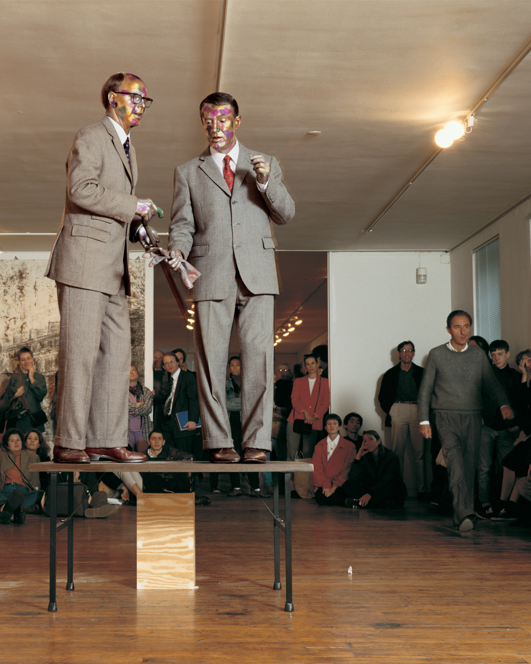 Two men in suits with painted faces stand on a table, performing before an audience in a gallery.