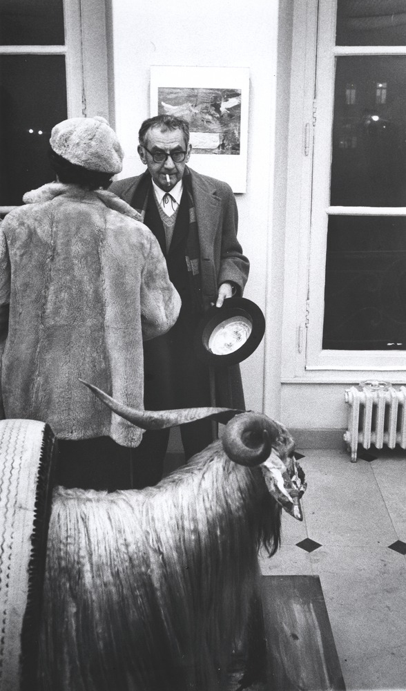 A man in a suit and overcoat smokes while talking to a woman near a taxidermied ram.
