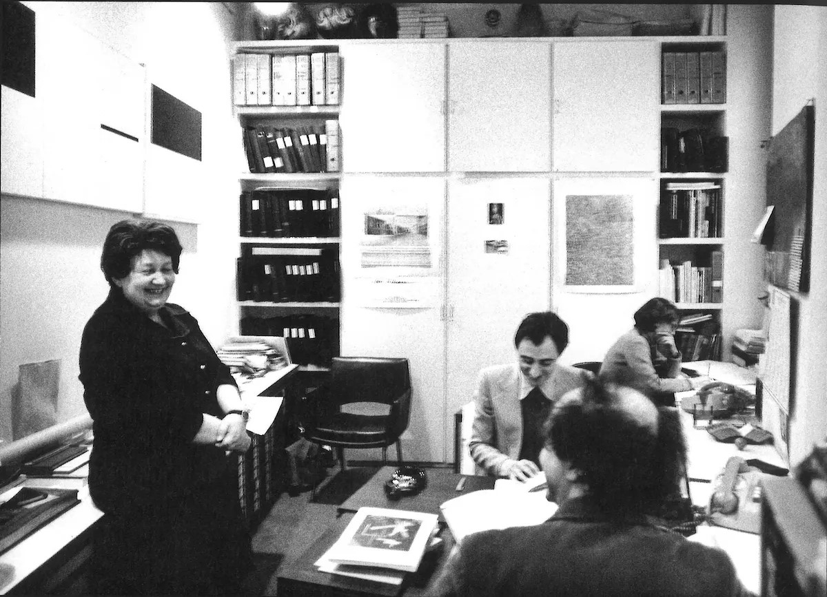 Four people are working and talking in a small, book-filled office.