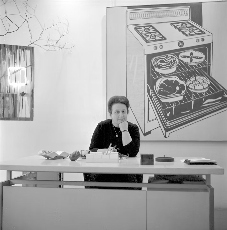 A woman sits at a desk in front of a large painting of a stove and oven with food inside.