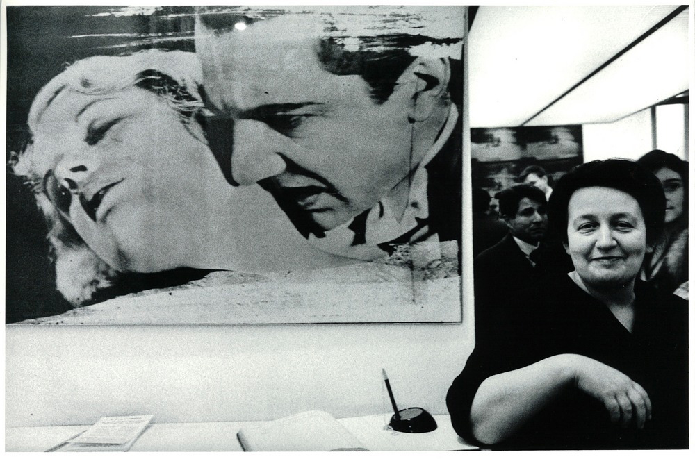 A woman stands smiling in front of a large, dramatic black-and-white artwork of two faces.