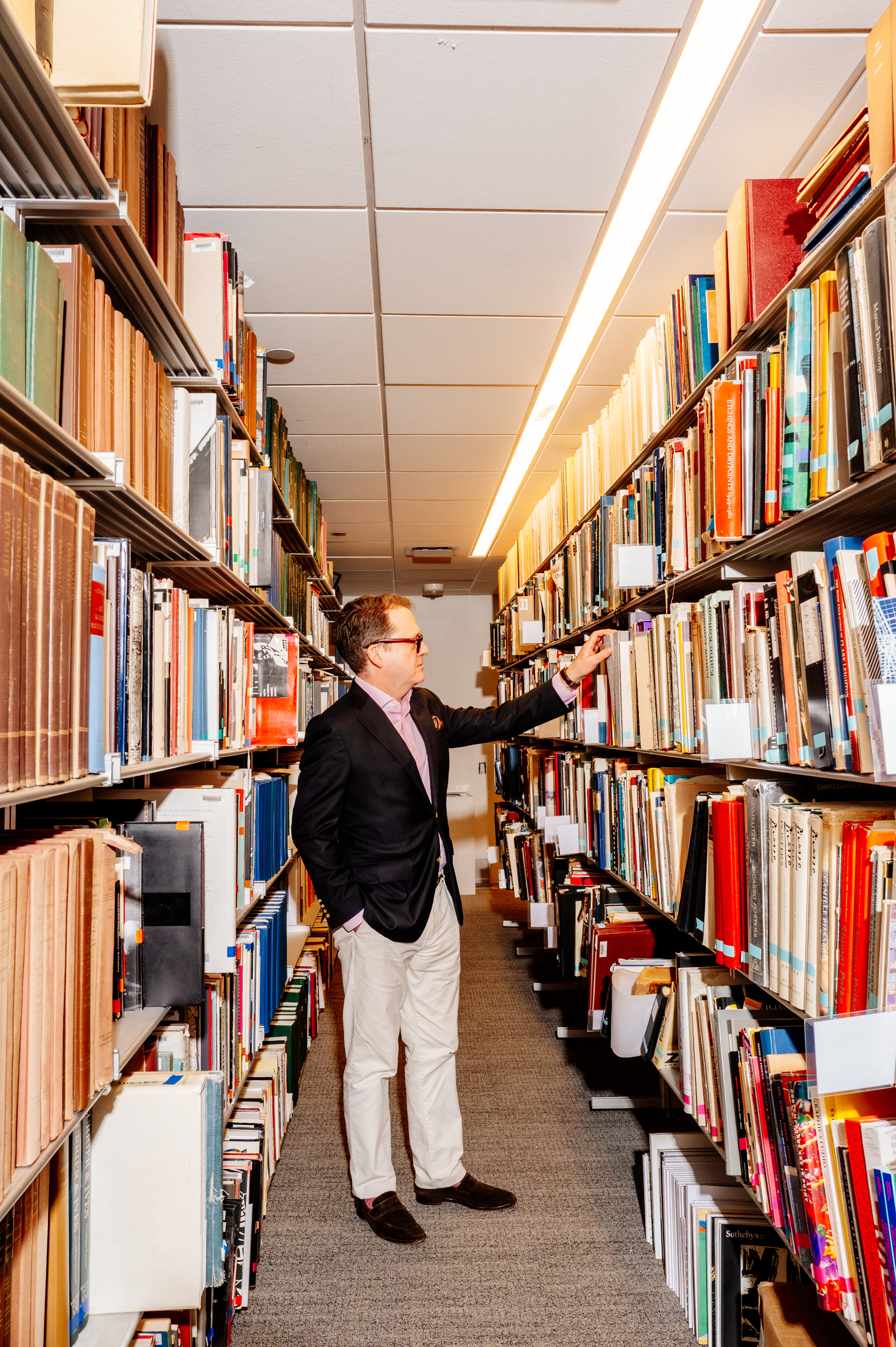 A man in a blazer is looking at books on a shelf in a library aisle.