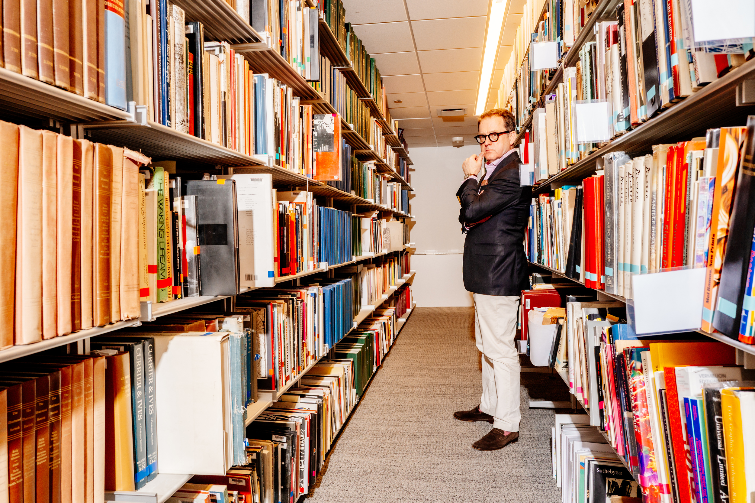 A man in a suit stands between bookshelves in a brightly lit library aisle.