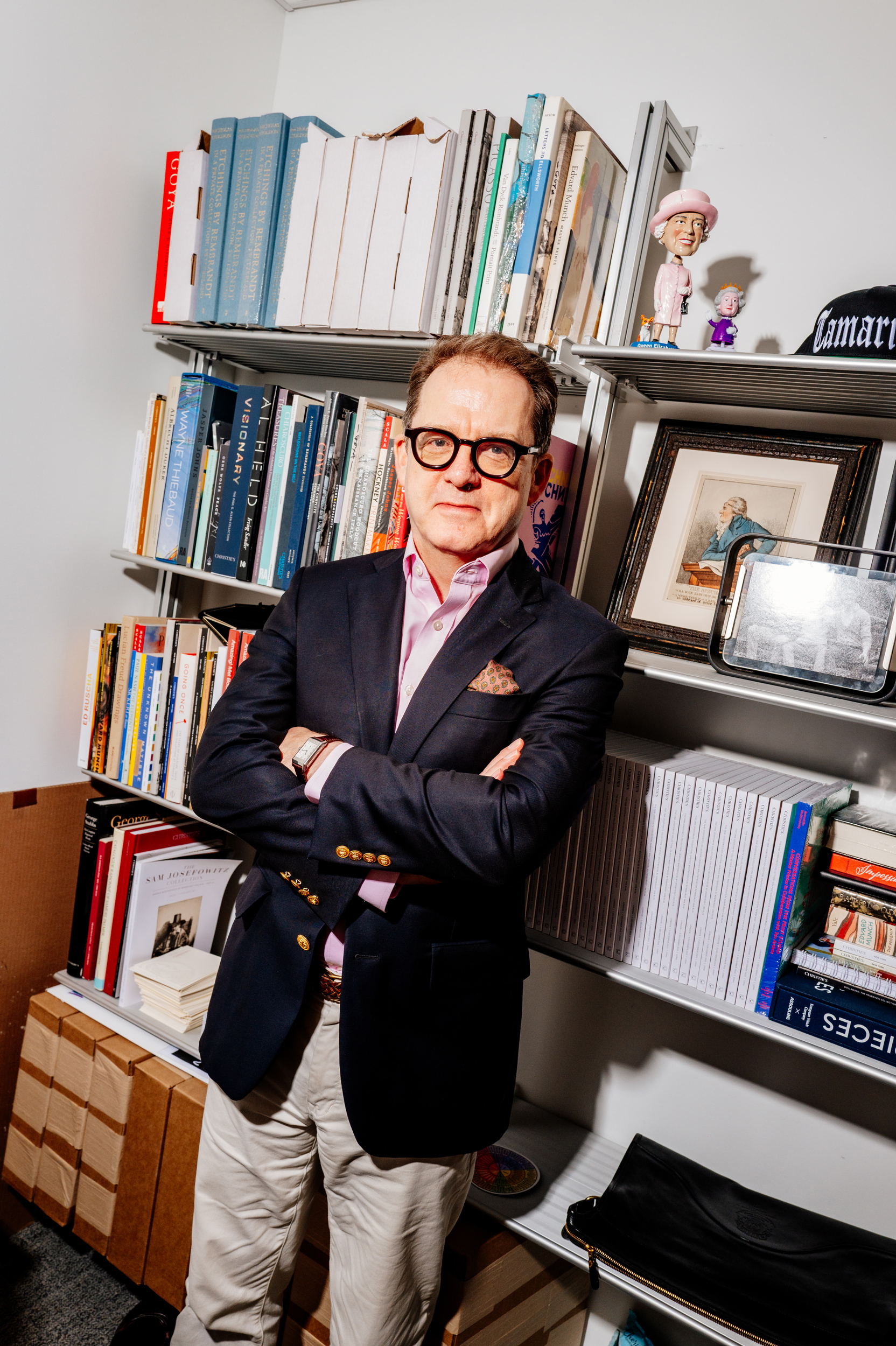 A man in glasses and a blazer stands with arms crossed in front of shelves filled with books.