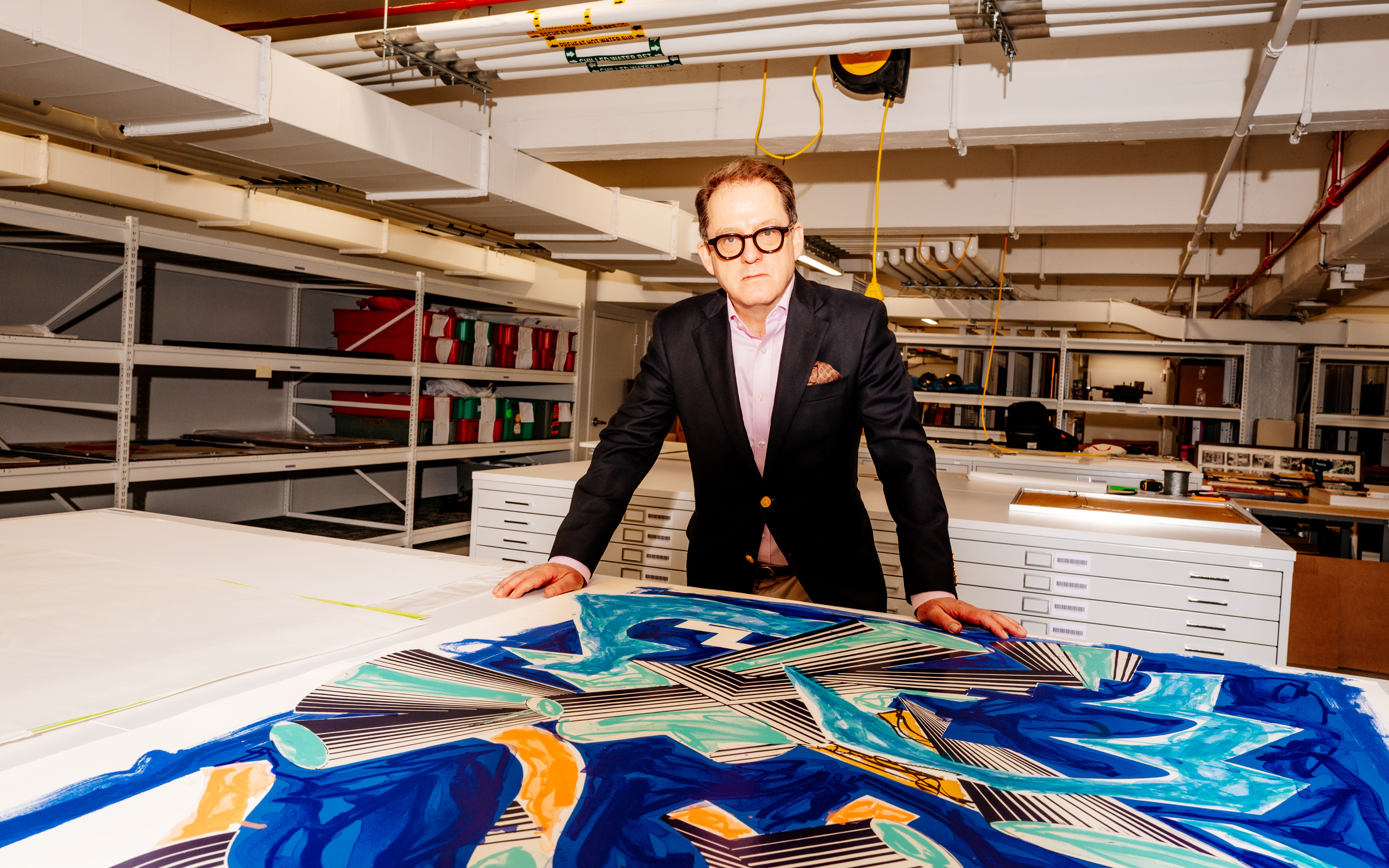 A man in a suit stands behind a colorful abstract artwork in an organized storage room.
