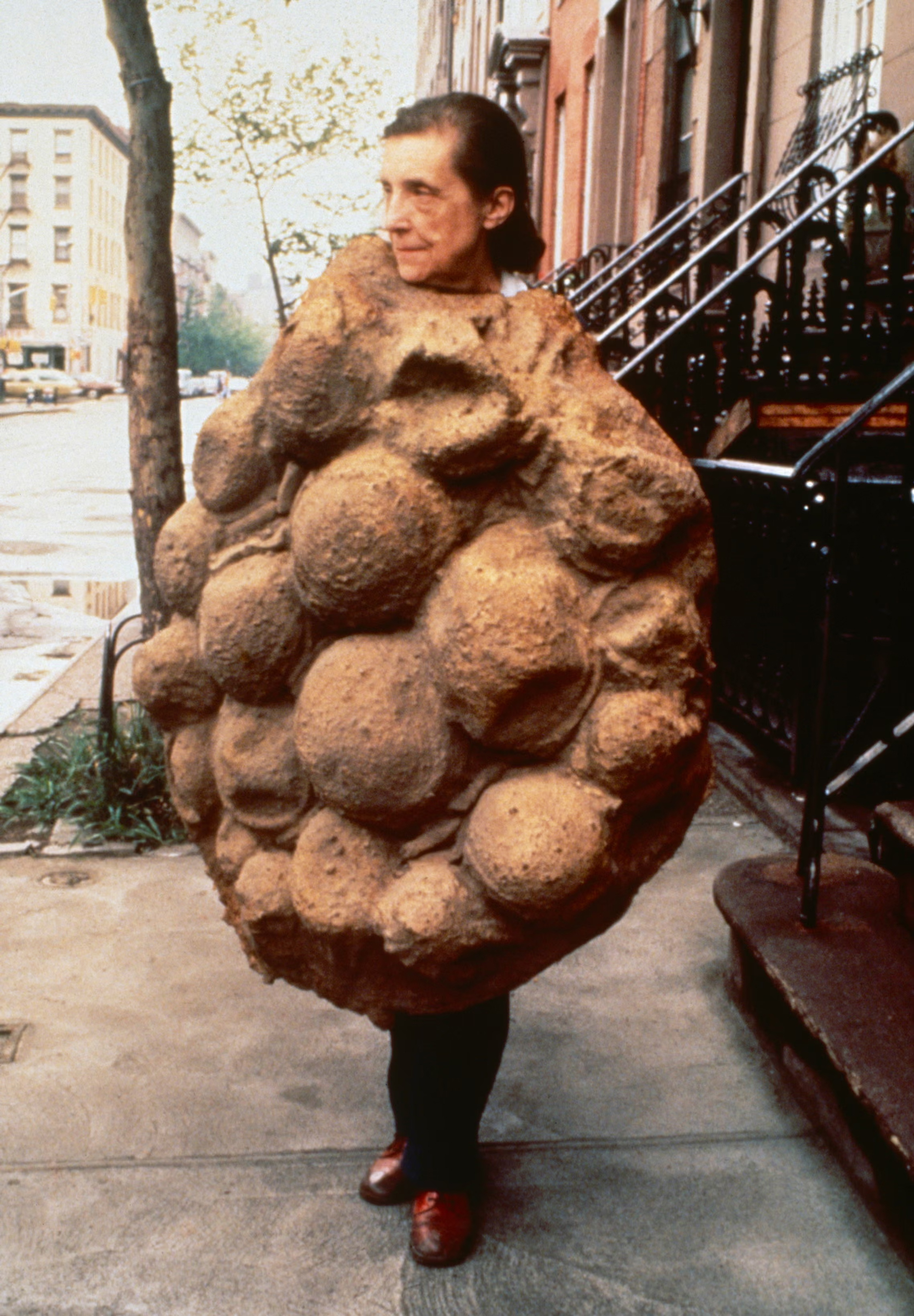A person is wearing a large, textured, brown, sculptural suit covered in round shapes on a sidewalk.