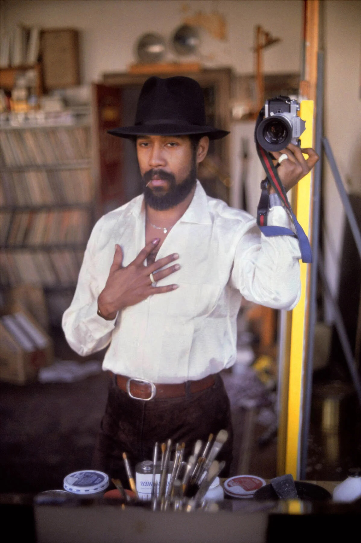 A man in a white shirt and black hat takes a mirror selfie in an art studio.
