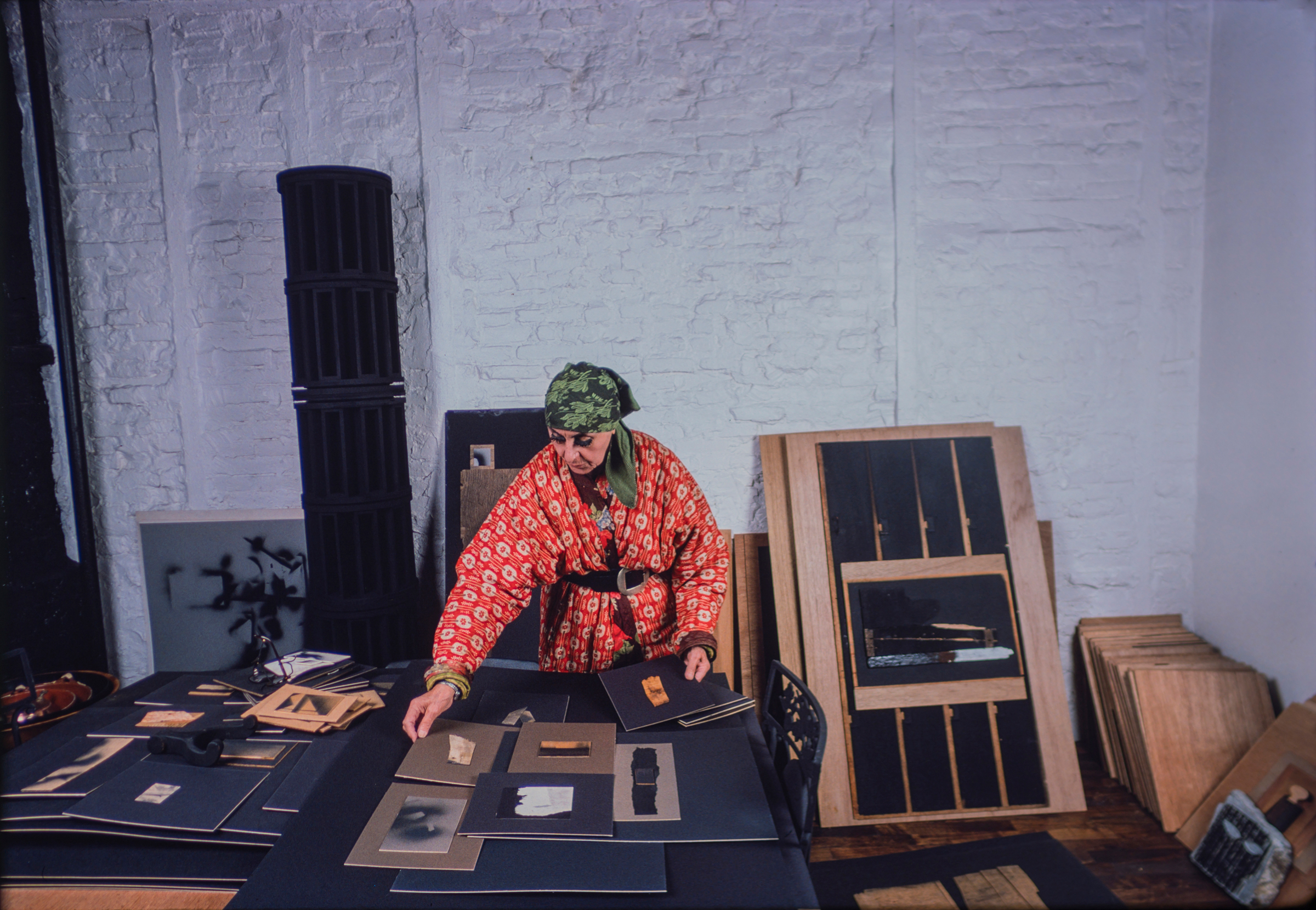 A person in a red patterned jacket arranges artwork on a table in a studio with white brick walls.