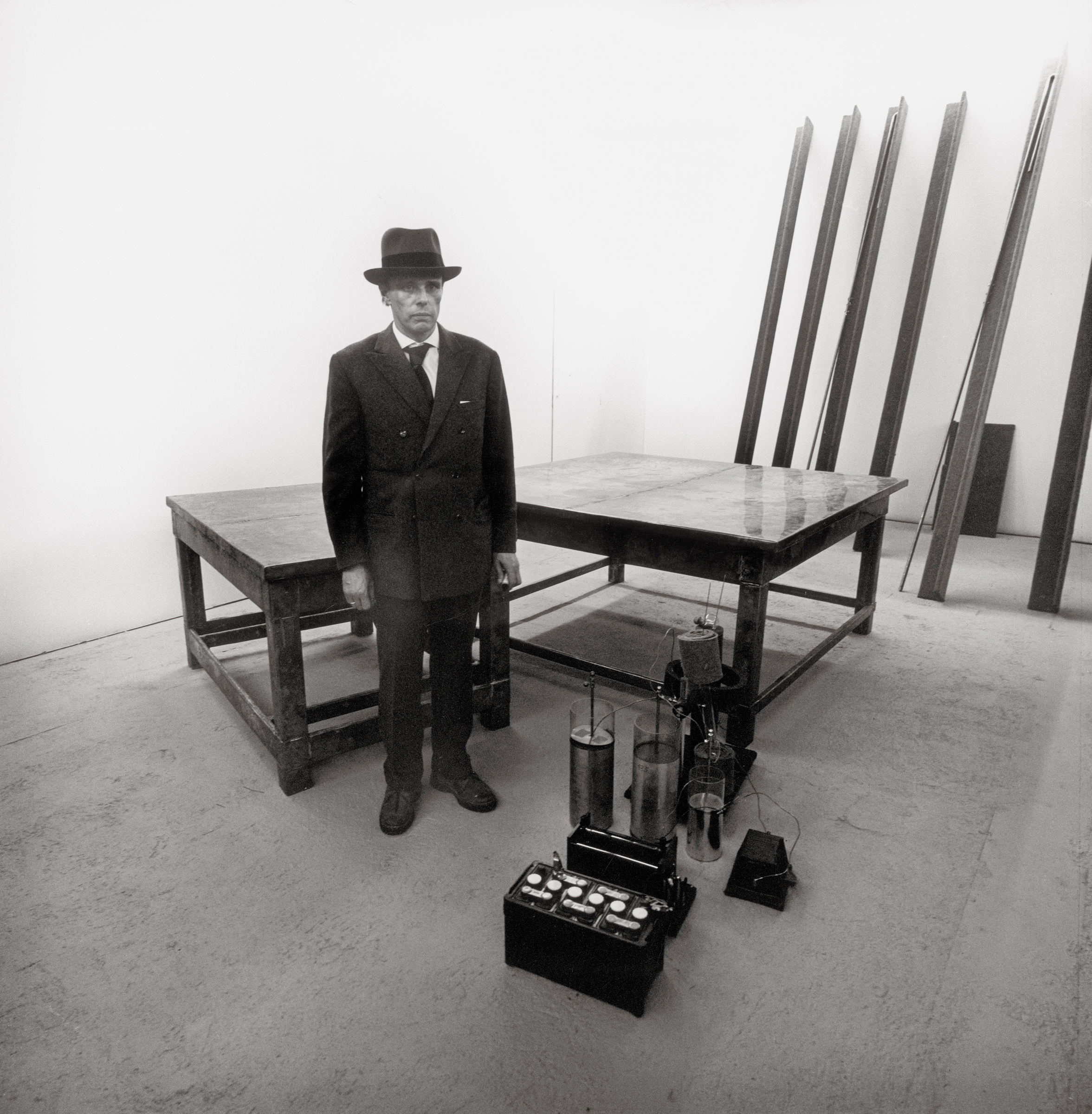 A man in a suit and hat stands in a sparse room with tables, metal rods, and electronic equipment.