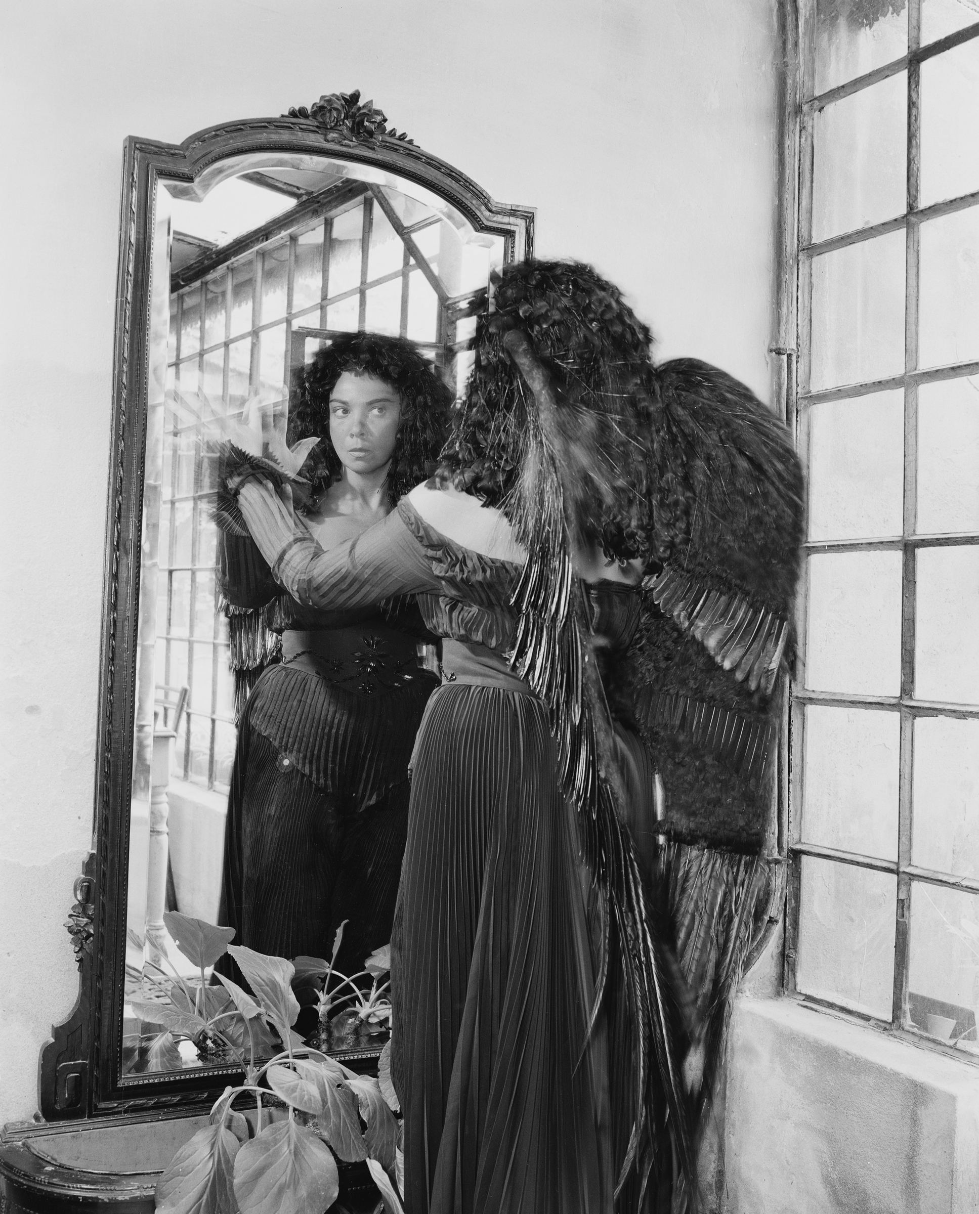 A woman with curly hair and angel wings looks at herself in a large ornate mirror.