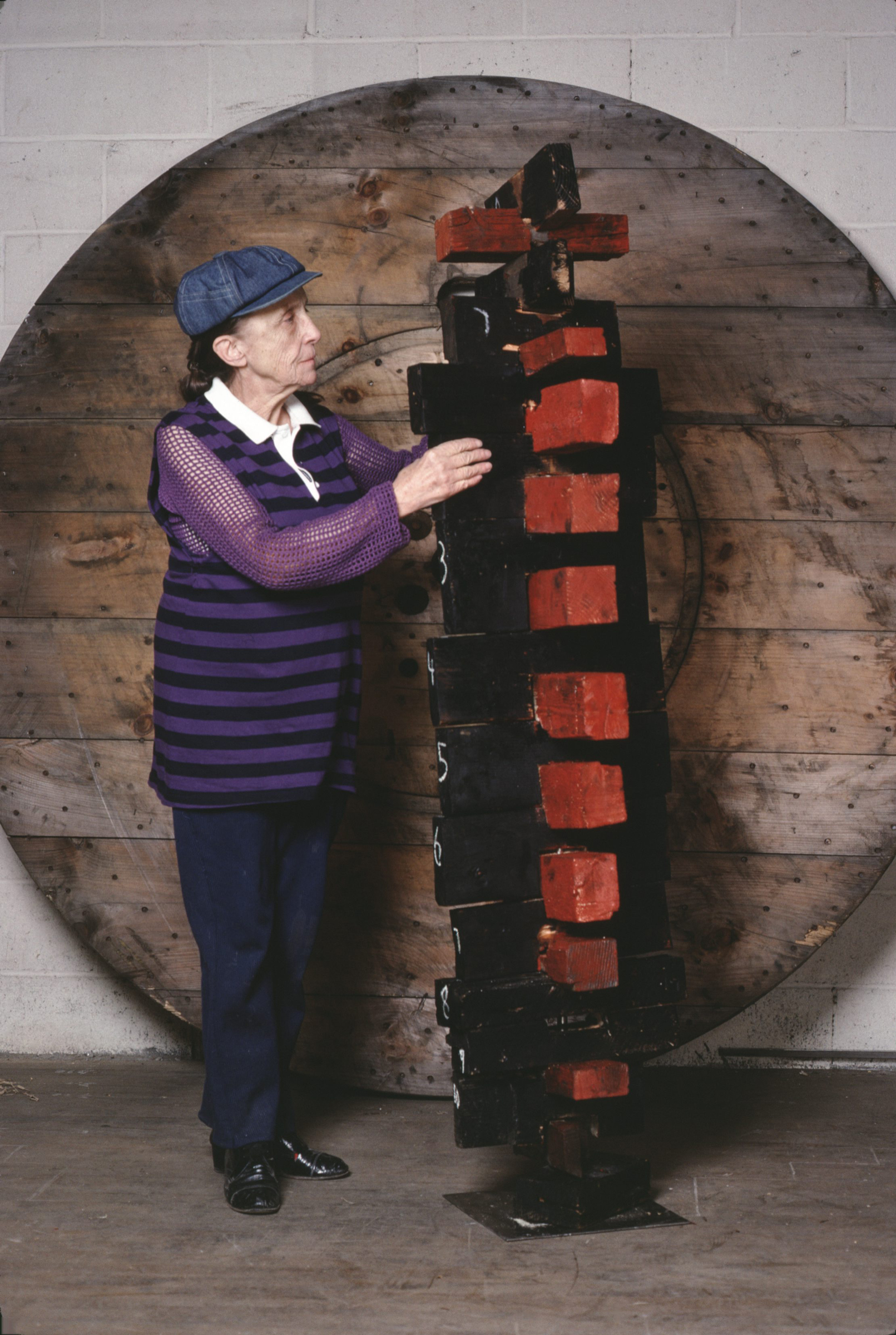 An elderly person wearing a cap stands beside a tall, abstract wooden sculpture indoors.