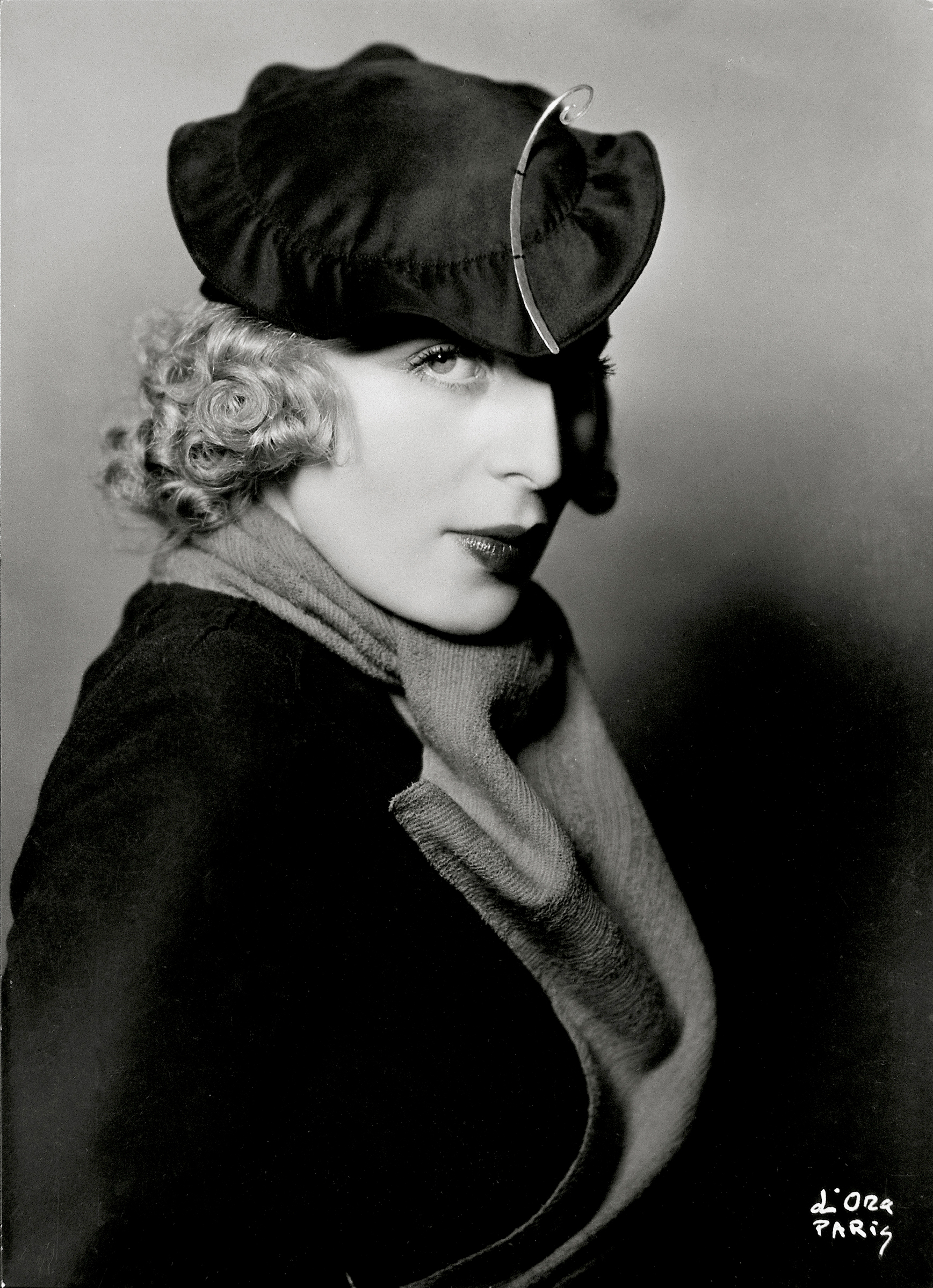 A woman wearing a stylish hat and scarf poses in a dramatic black and white portrait.