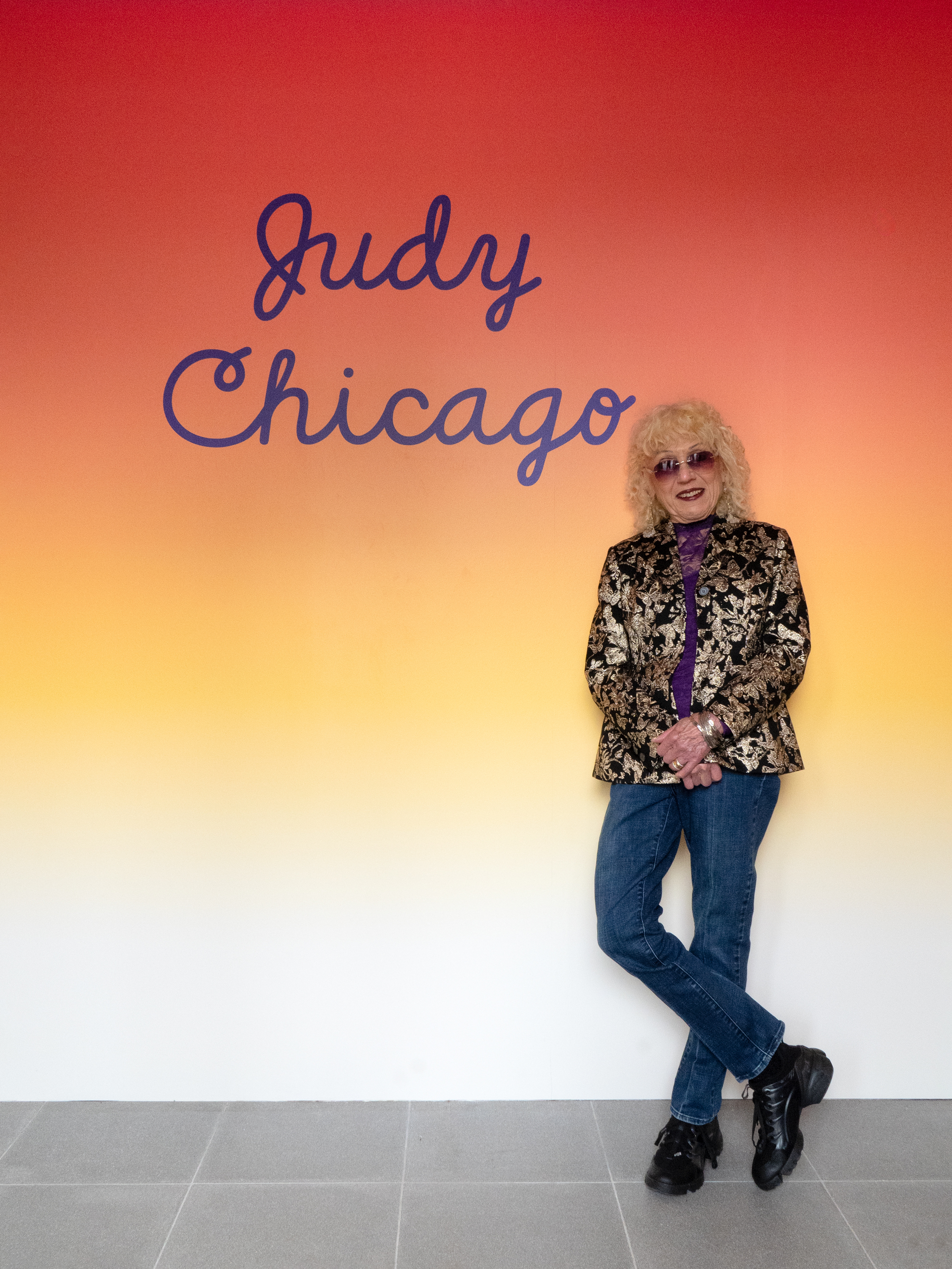 A person with curly blonde hair stands in front of a wall with "Judy Chicago" written on it.