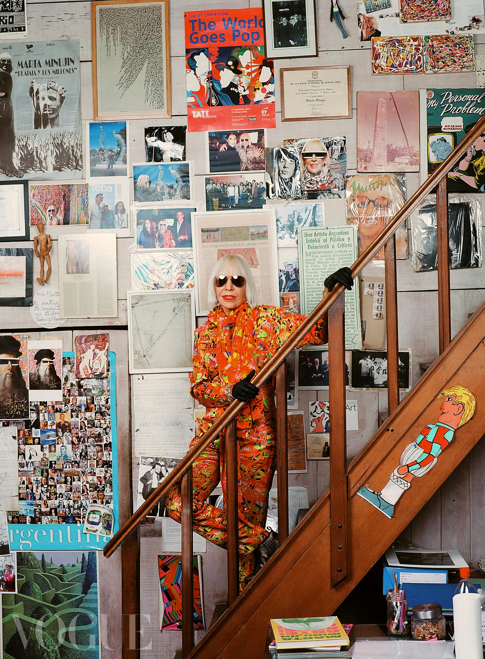 A woman in a colorful outfit stands on stairs surrounded by a wall covered in photos and art.