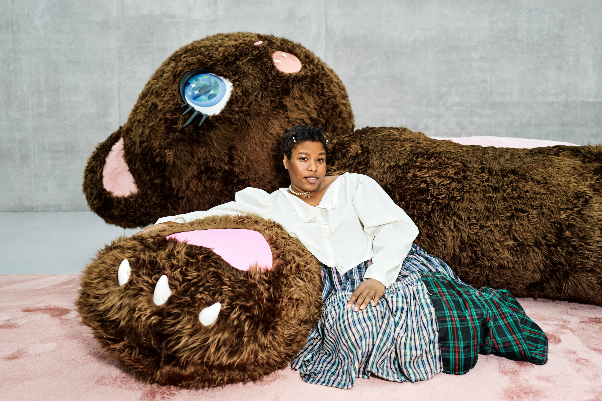 A person in a white blouse and plaid skirt leans against a giant plush bear on a pink floor.