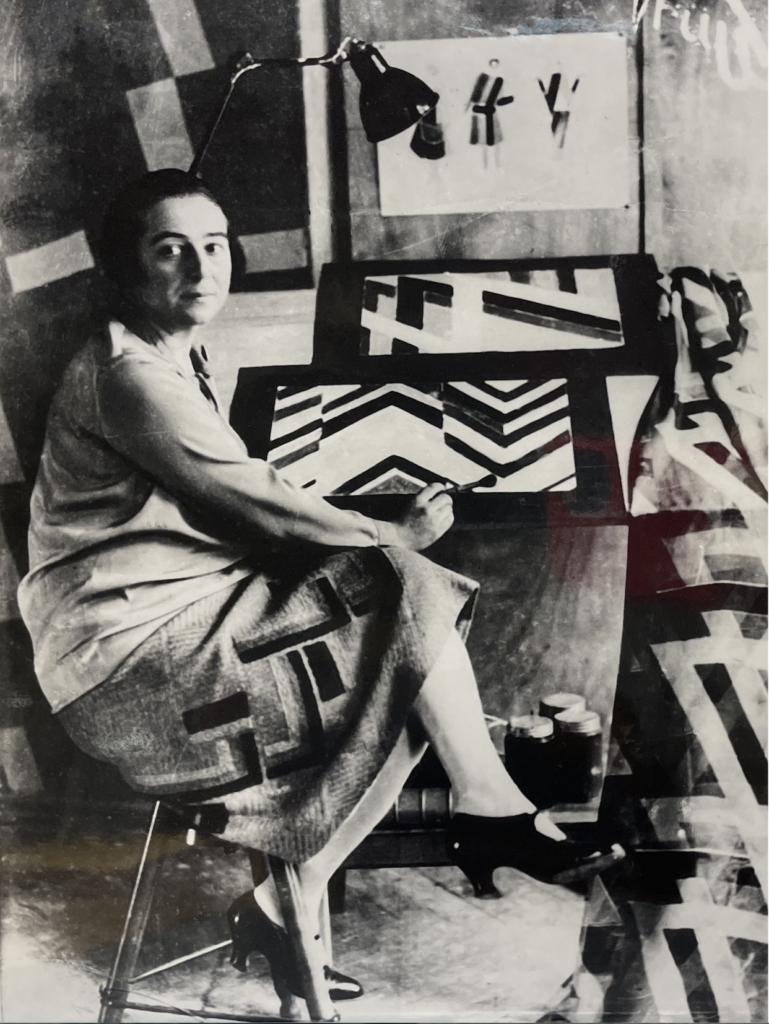 A woman sits at a desk painting geometric patterns, with art supplies and abstract art around her.