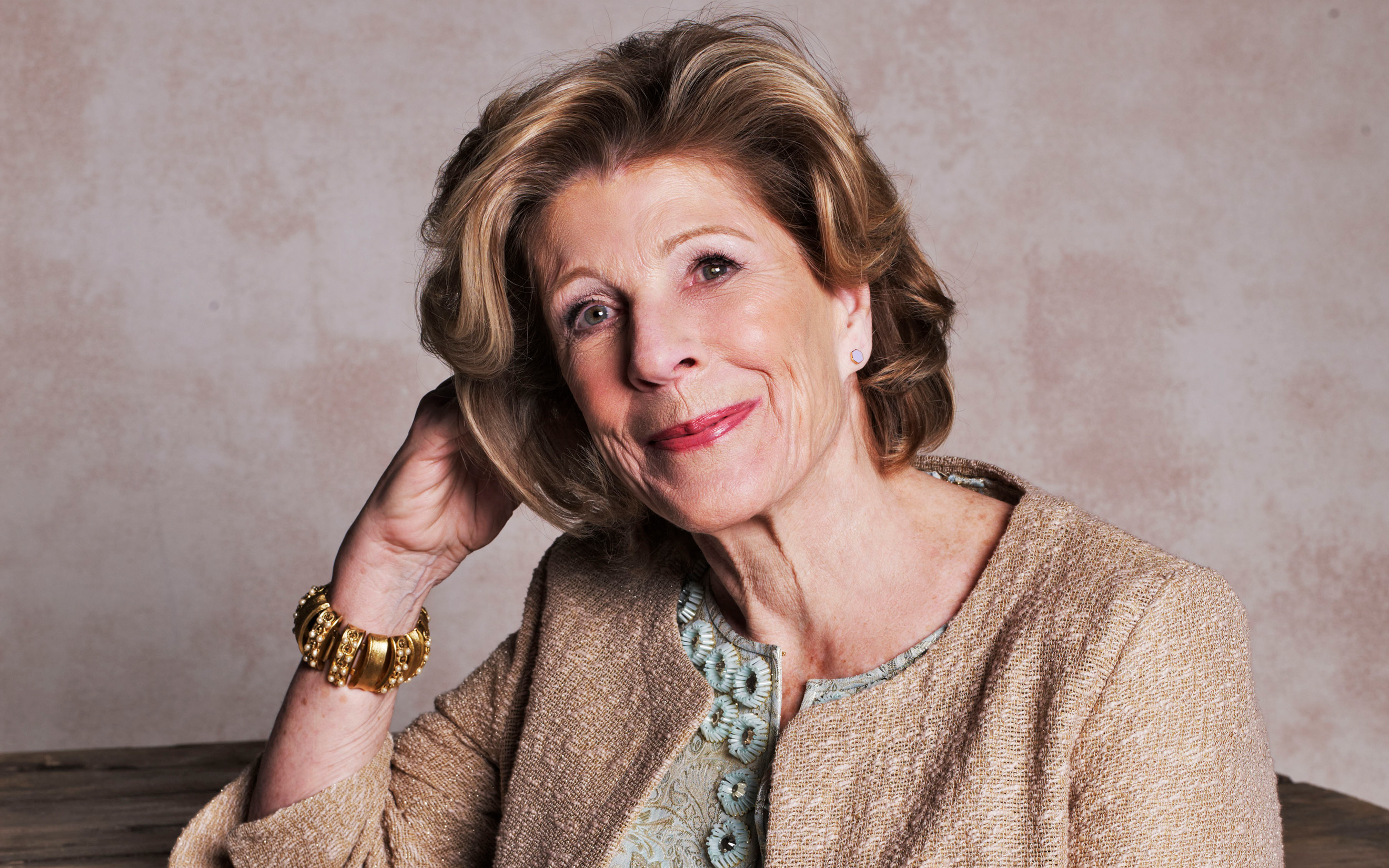 An older woman with styled hair and a gold jacket is smiling and resting her head on her hand.