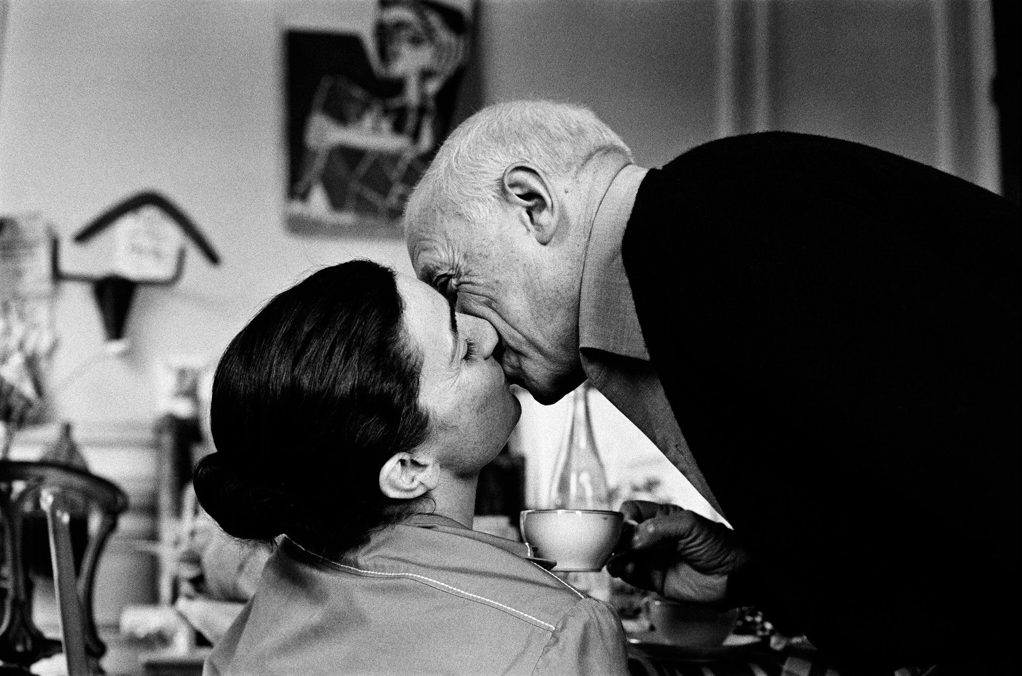An elderly man and a woman share a kiss in a cozy indoor setting with art on the wall.