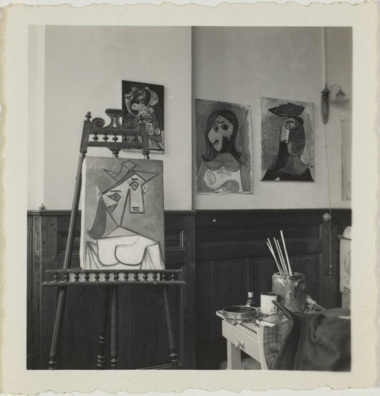 The image shows an artist's studio with abstract paintings on the wall and an easel with a canvas.