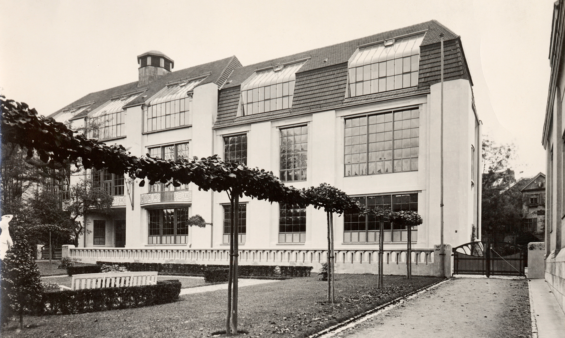 A large building with many windows stands behind a landscaped garden and a row of trimmed trees.