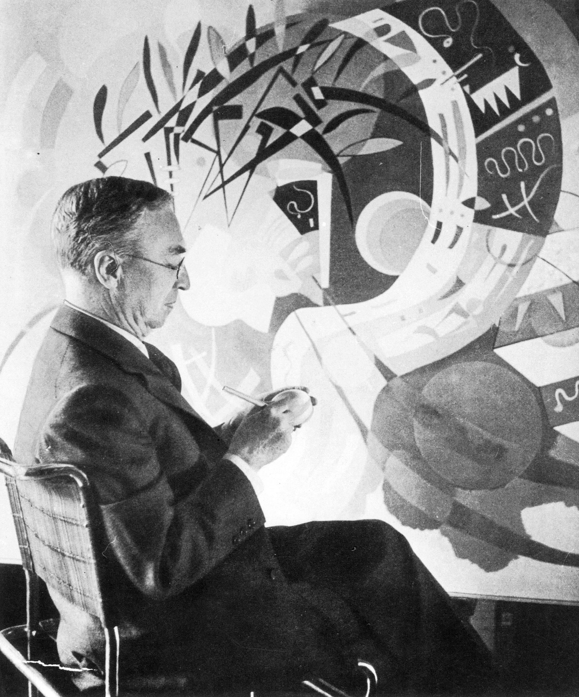 A man in a suit sits and sketches in front of an abstract, geometric mural.