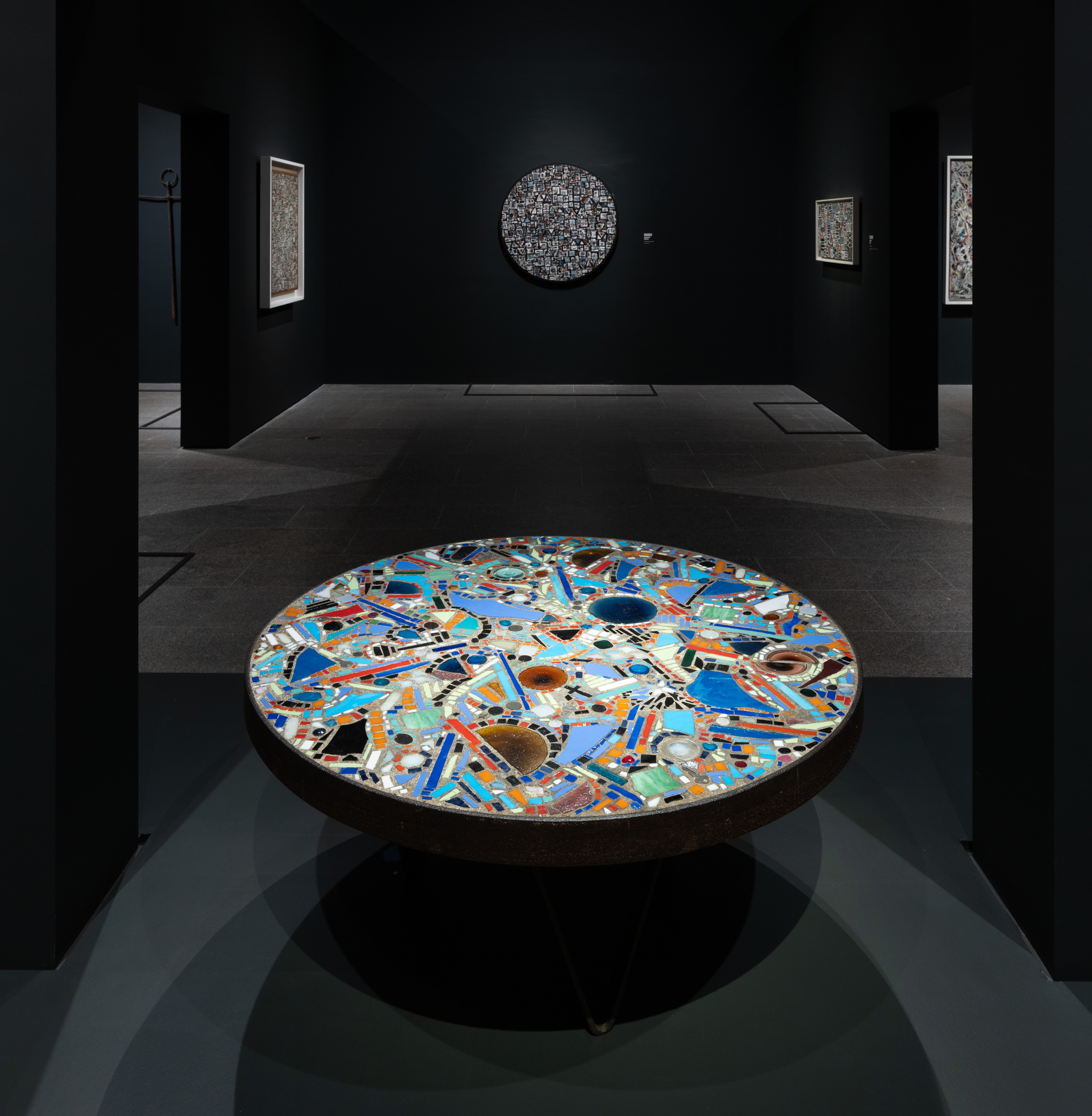 A round mosaic table with colorful patterns is displayed in a dark, modern art gallery.