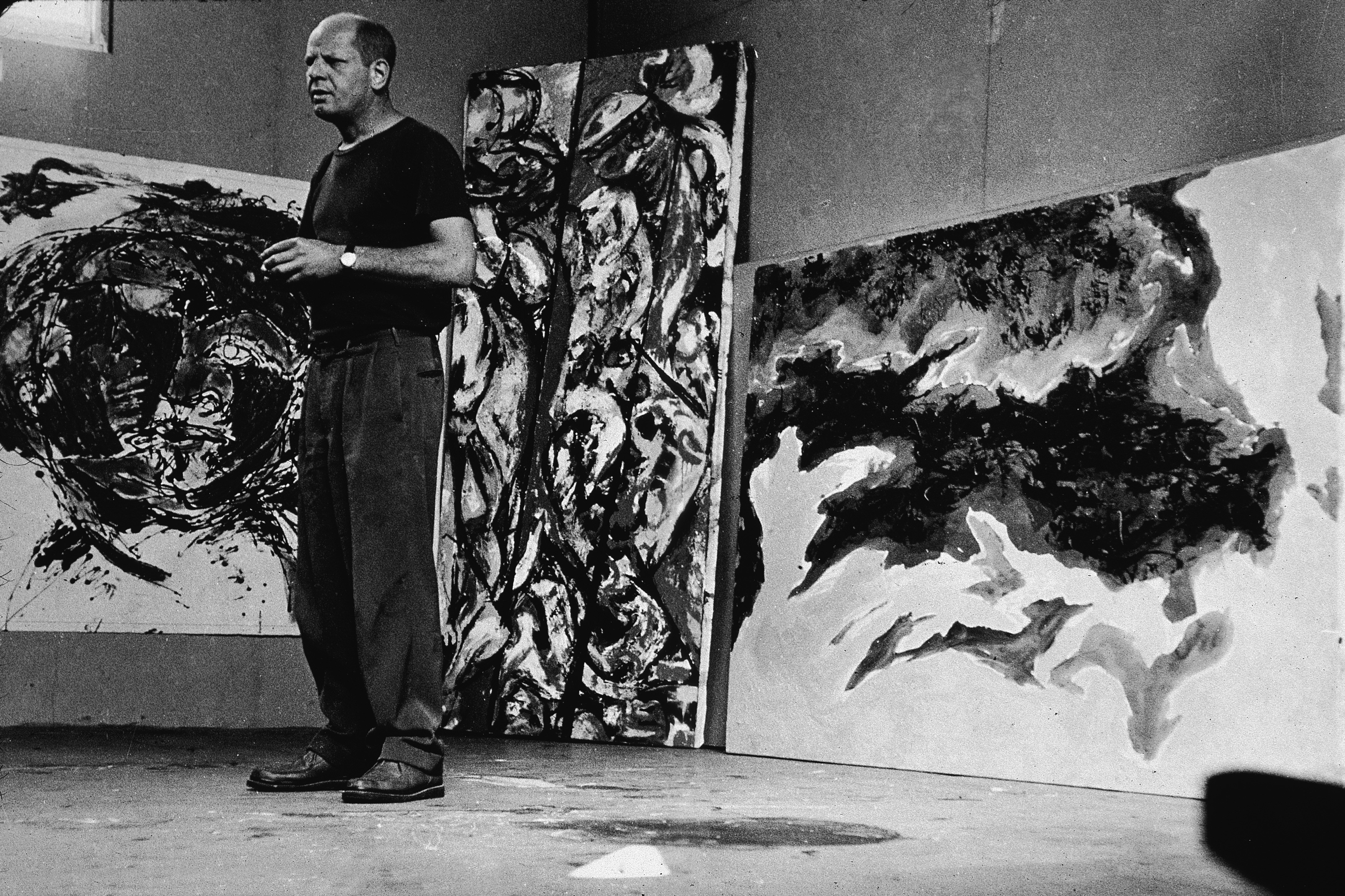 A man stands in front of large abstract paintings in an art studio.