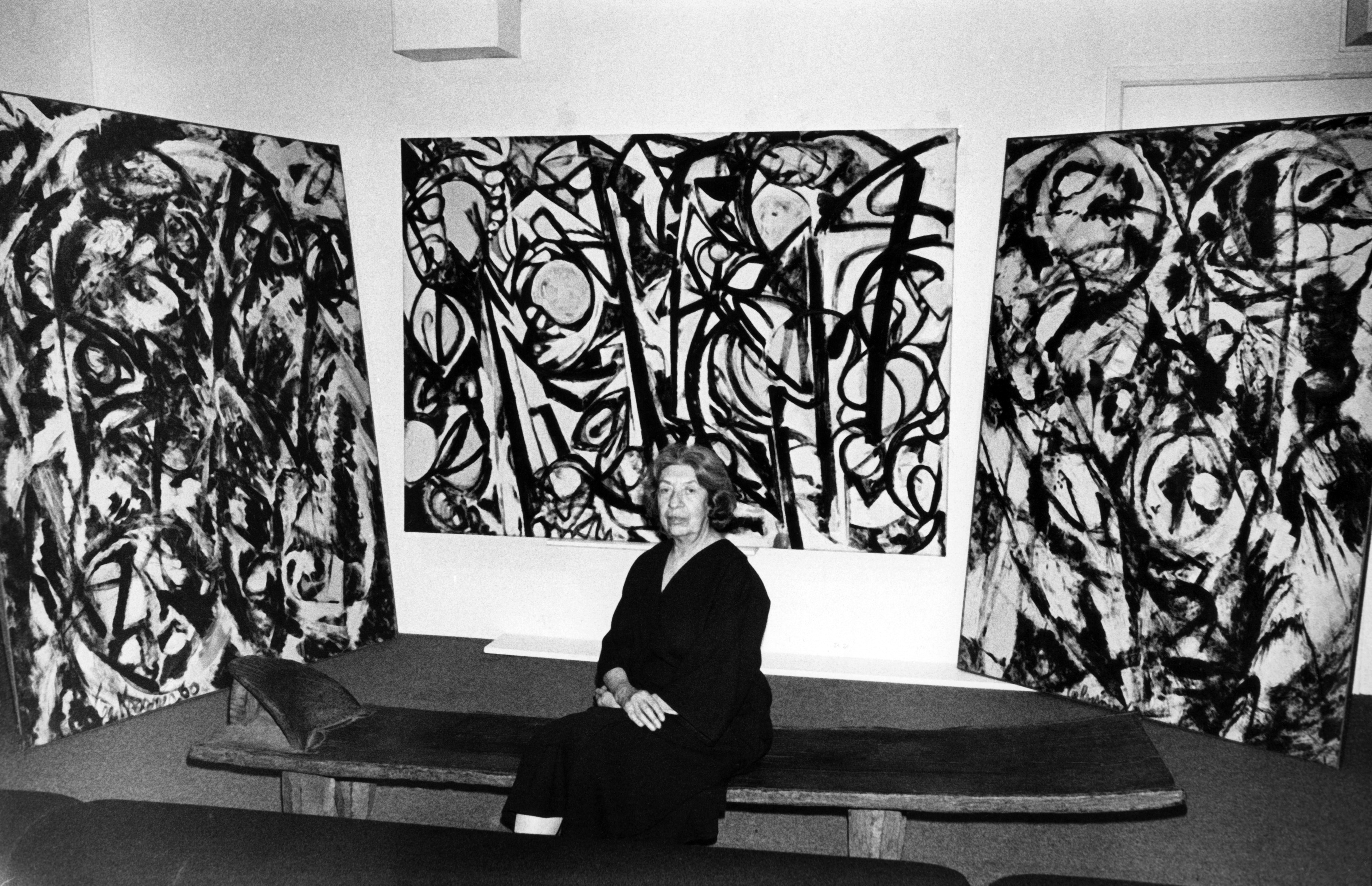 A woman sits on a bench in front of three large abstract black-and-white paintings.
