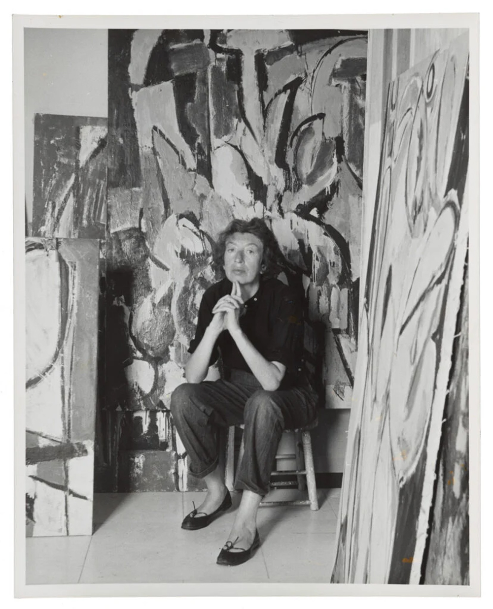 A woman sits on a stool surrounded by large abstract paintings in a studio.