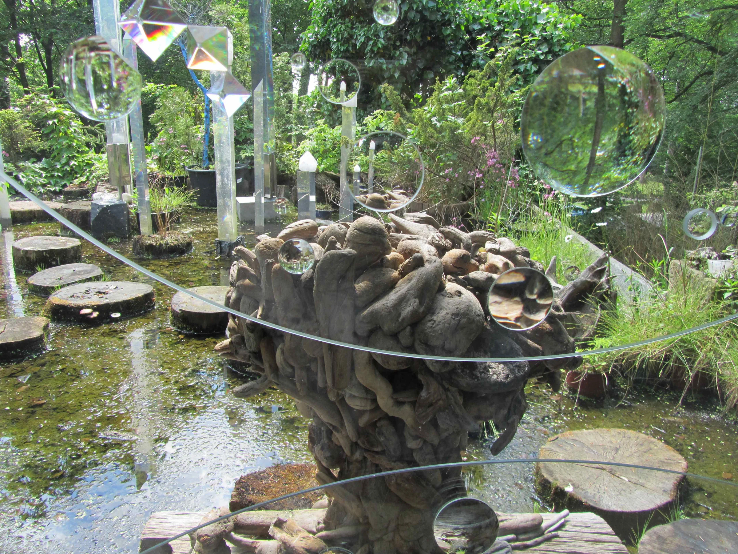 A wooden sculpture stands in a garden pond surrounded by glass orbs and reflective structures.