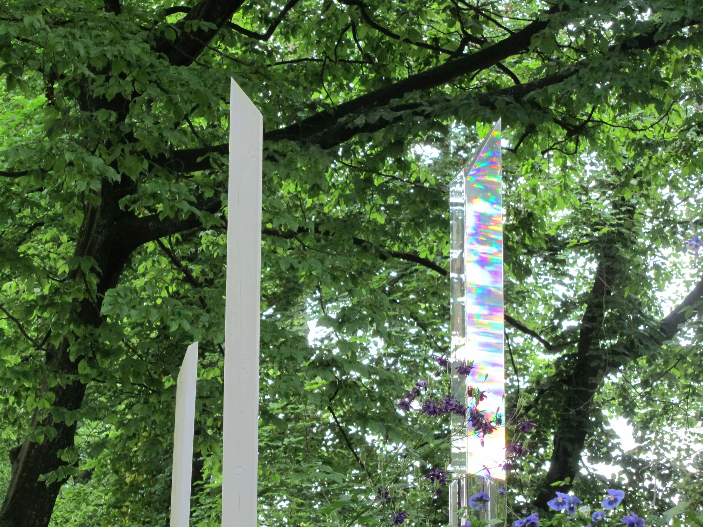 Three tall, slender sculptures stand among green trees and purple flowers, one reflecting rainbow colors.