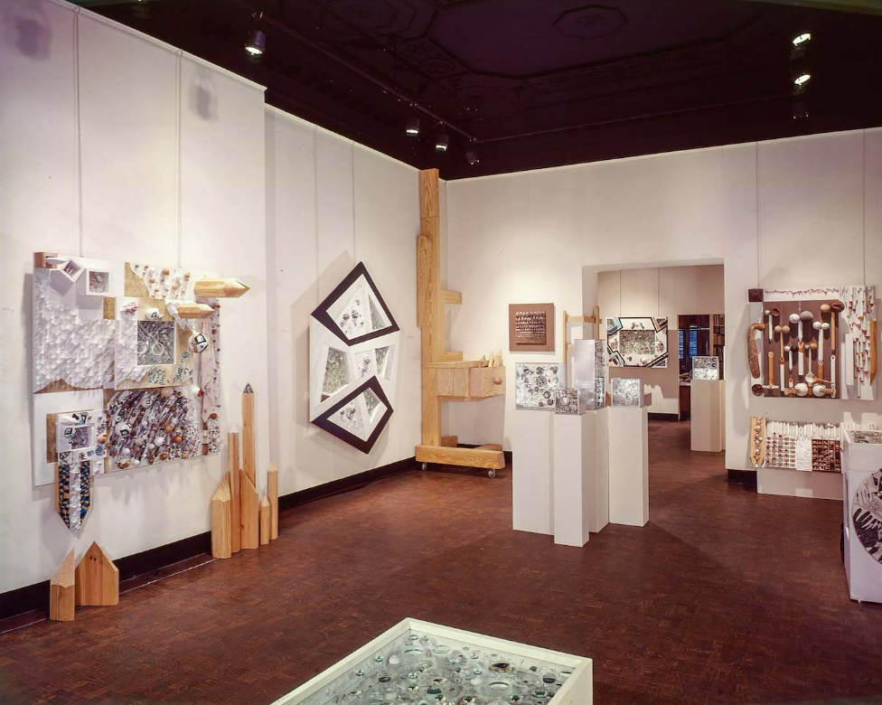 An art gallery room displays various abstract mixed-media artworks on white walls and pedestals.