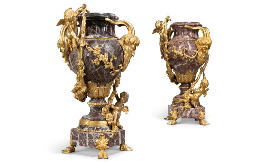 Two ornate marble vases with gold figurative and floral embellishments, each supported by a small cherubic figure.