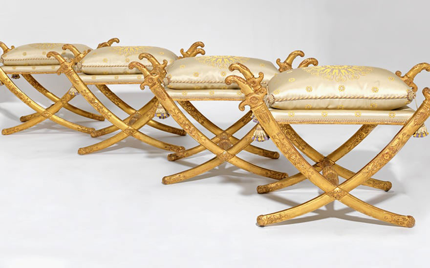 Four ornate golden folding stools with white and gold embroidered cushions displayed in a row.