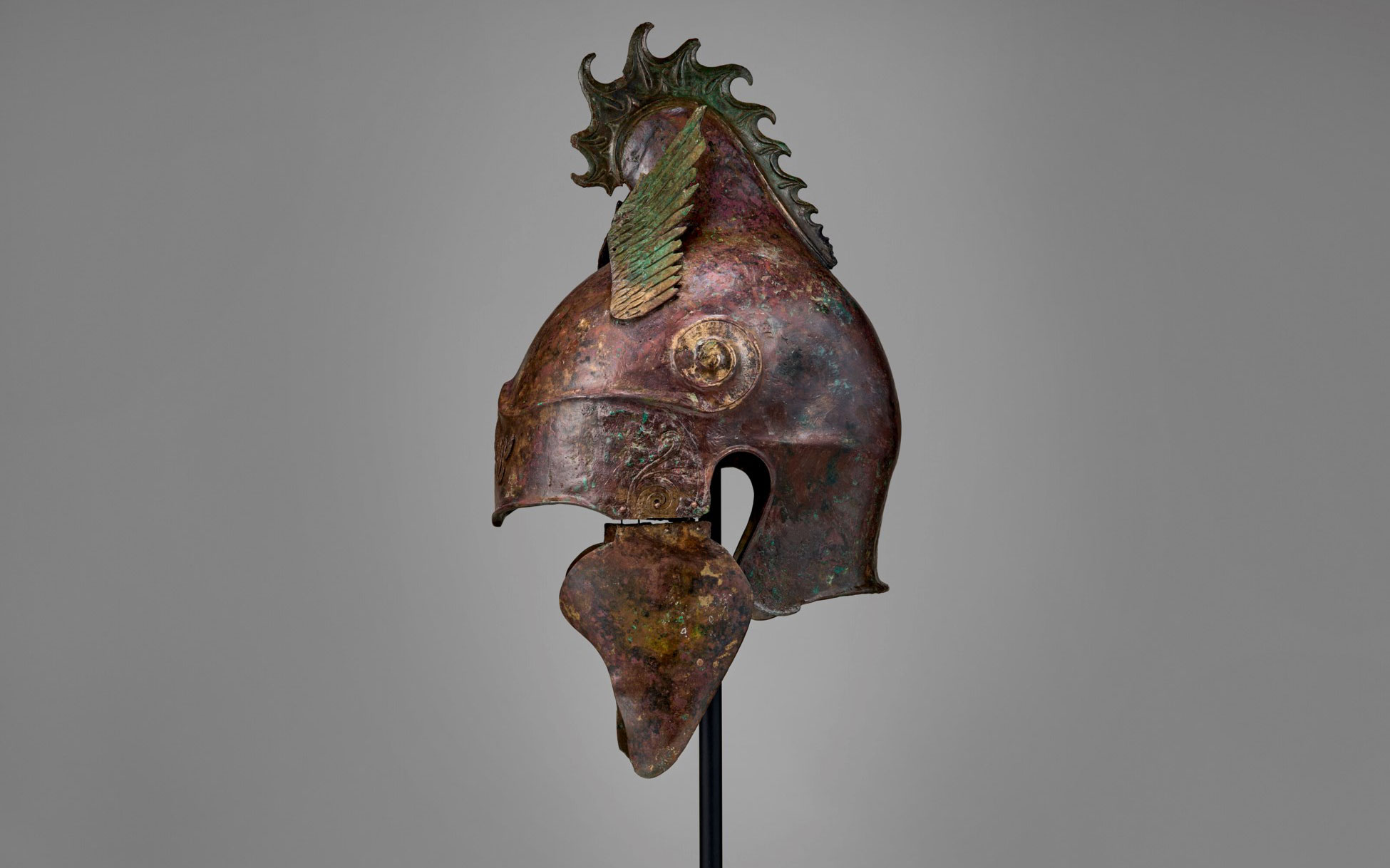 An ancient bronze helmet with a decorative crest, possibly of Greek or Roman origin, displayed on a stand.