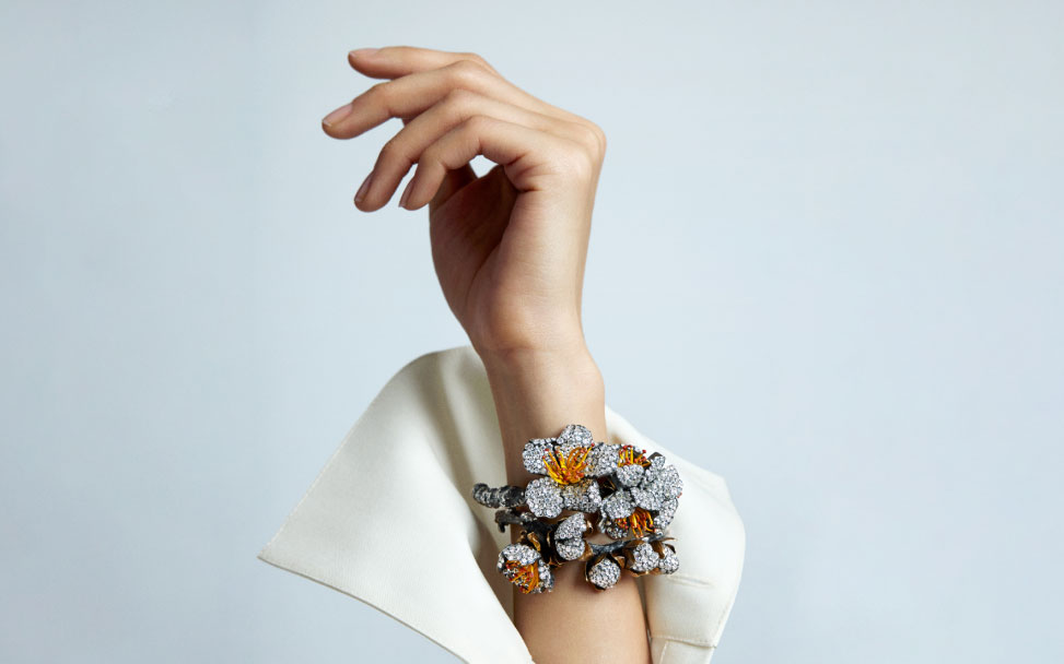 A woman's hand adorned with a luxurious floral bracelet featuring diamonds and colorful gemstones, poised elegantly against a light background.