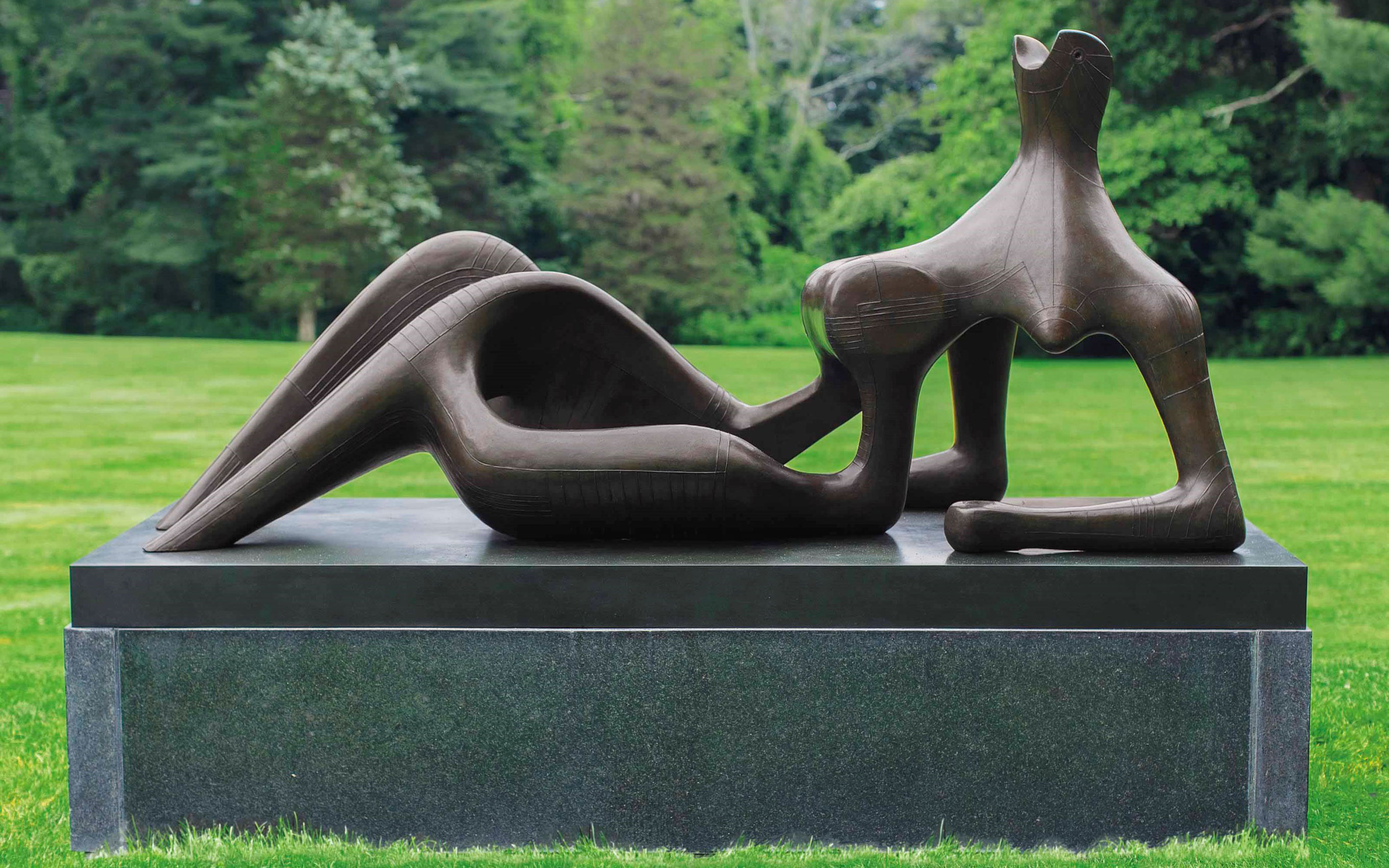 A bronze sculpture depicting a reclining female figure and a sitting animal figure, set on a pedestal in a grassy field.
