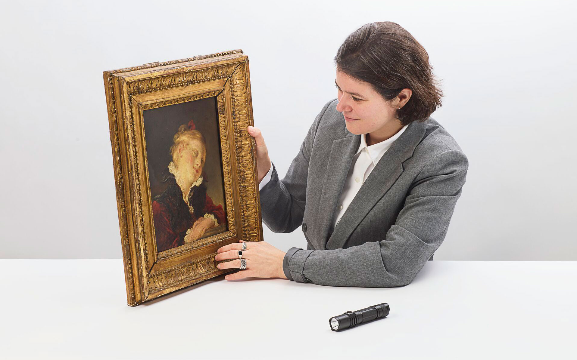 A woman in a gray suit examines a framed painting while sitting at a white table.