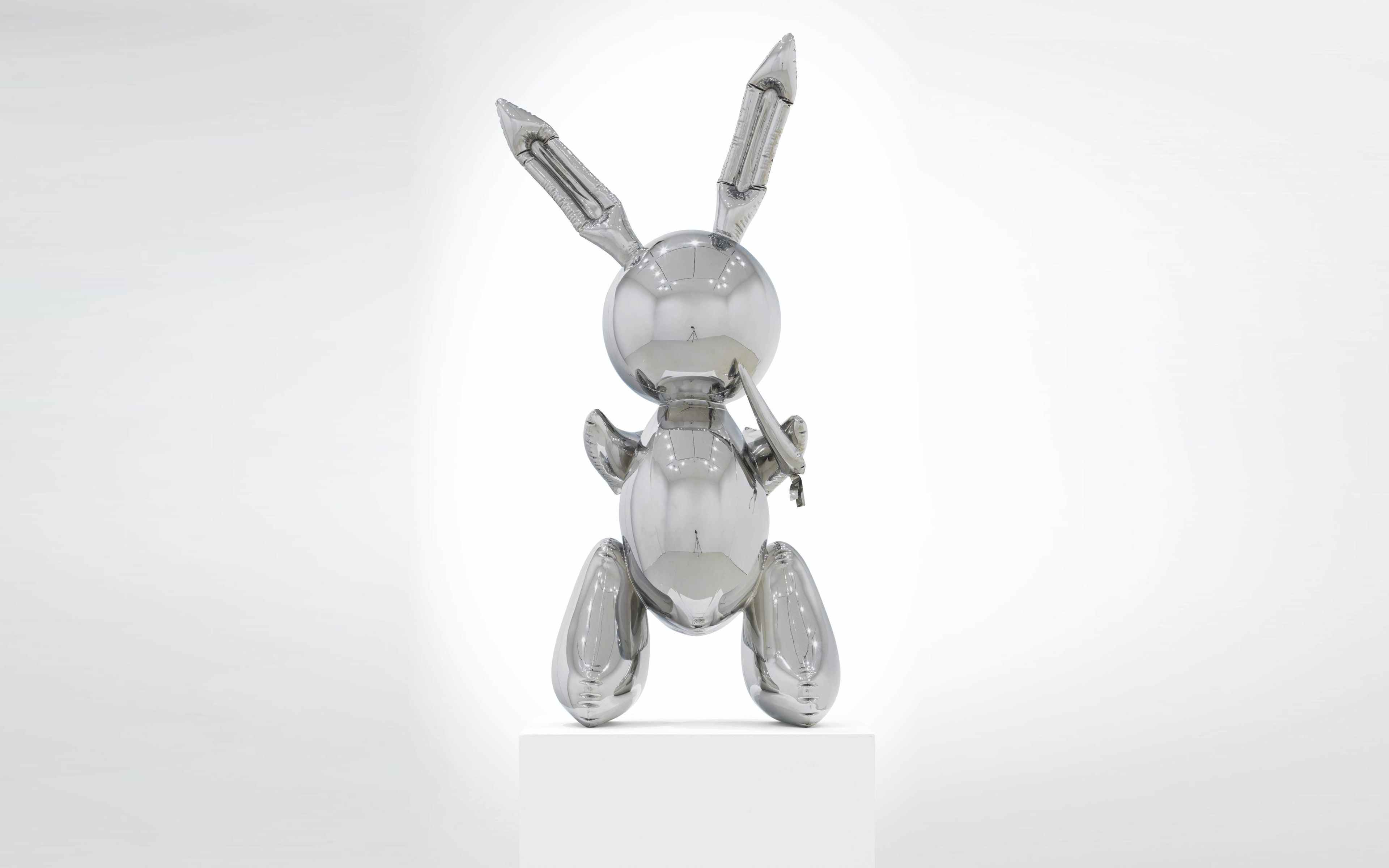 A reflective, metallic sculpture of a rabbit, shown in a sitting pose on a white pedestal against a gradient background.
