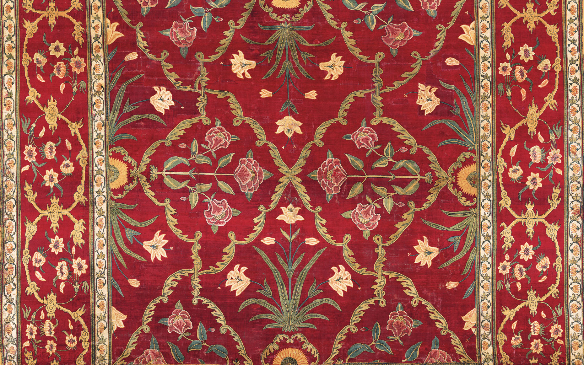 An ornate traditional rug featuring intricate floral and vine patterns in gold and beige on a deep red background.