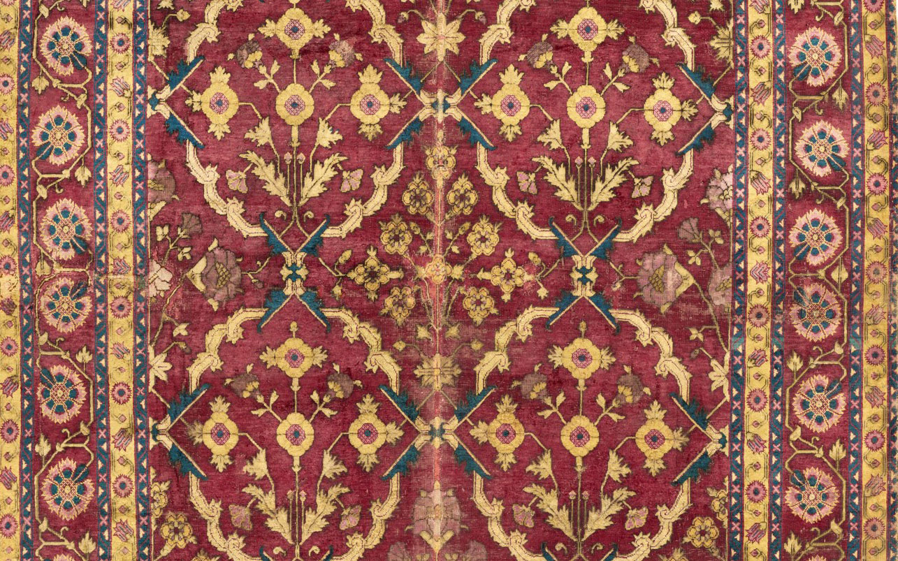 Detailed view of a traditional ornate carpet featuring symmetrical floral and geometric patterns in rich maroon, blue, and gold tones.