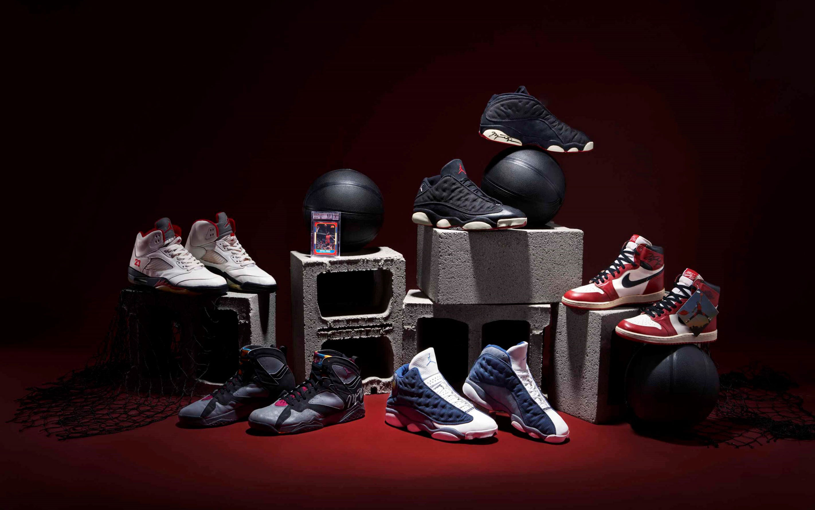 Assorted basketball sneakers are displayed artistically on concrete blocks and spheres against a dark red background.