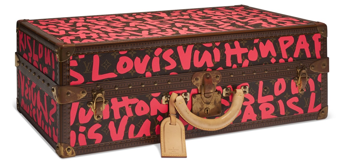 A Louis Vuitton trunk with bold red graffiti-style lettering on a brown monogram background.