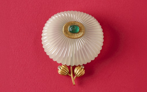 A white and gold flower-shaped brooch with a green gemstone center on a red background.