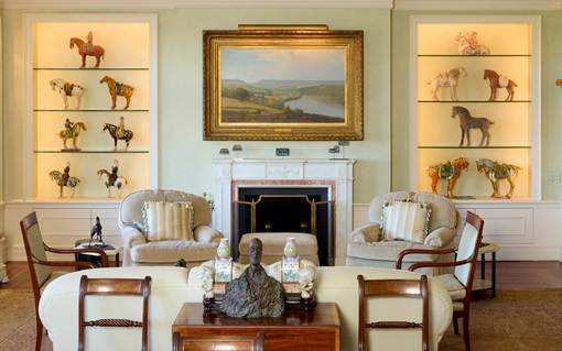 A traditional living room with armchairs, display shelves of horse figurines, and a fireplace.