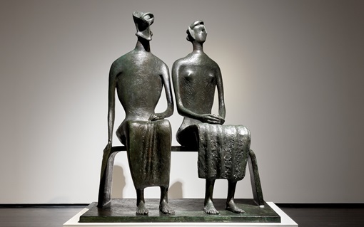 A bronze sculpture of two abstract human figures seated closely together on a bench.
