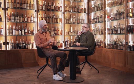 Two people are sitting at a round table, tasting drinks in a room lined with shelves of liquor bottles.