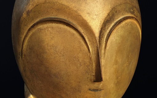 A close-up of a smooth, golden abstract sculpture resembling a simplified human face.